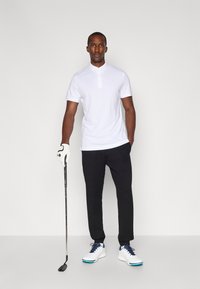 Man i vit polo och svarta byxor står med en golfklubba i handen, med en vit handske på vänster hand, framför en enkel bakgrund.
