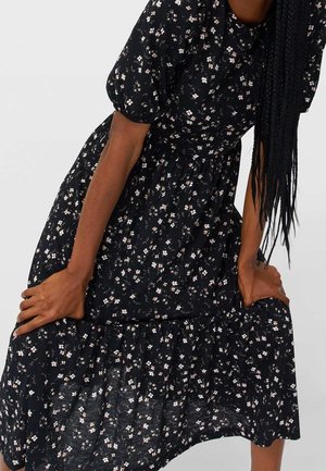 Personne aux longs cheveux tressés portant une robe noire à fleurs avec des manches courtes bouffantes, tenant l’ourlet de la robe.