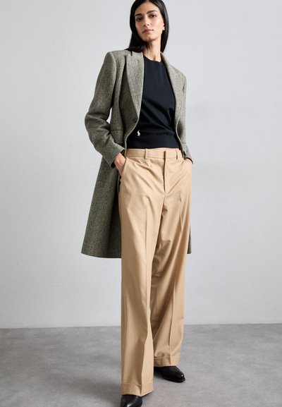 Ein graues und schwarzes Hahnentrittmuster-Mantel, kombiniert mit einem schwarzen Oberteil und beigen weiten Hosen, abgerundet mit schwarzen Schuhen.