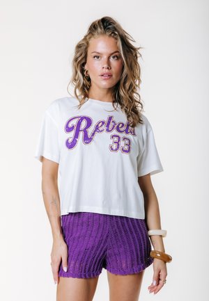 Femme aux cheveux blonds ondulés portant un T-shirt blanc "Rebels 33" et un short en maille violet, accessoirisée avec des bracelets et des bagues, debout sur fond blanc.