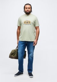 Άνδρας με πλεξουδάκια, φορώντας ανοιχτοπράσινο T-shirt με γραφικό αποτύπωμα κρανίου ηλίανθου, μπλε τζιν, μαύρα αθλητικά παπούτσια, κρατώντας μια ολιβ πράσινη τσάντα duffle.