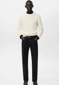 Uomo con maglione crema a coste sopra camicia nera, pantaloni neri e scarpe nere, in piedi con le mani nelle tasche davanti a uno sfondo semplice.