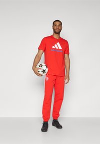 Røde sportsklær inkludert t-skjorte med hvit adidas-logo og teksten "BAYERN MÜNCHEN"; matchende røde bukser med hvite side-striper. Holder en fotball.
