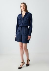 Camicia di lino navy con maniche lunghe e due tasche sul petto, abbinata a pantaloni corti su misura dello stesso colore, completata da sandali con tacco marroni.