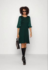 Vestido verde con mangas abullonadas de lunares, dobladillo acampanado, combinado con botas negras altas hasta la rodilla de charol y un pequeño bolso negro.