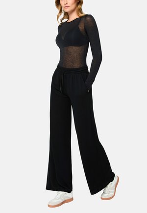 Kvinde iført en gennemsigtig sort langærmet top, sorte vidde bukser med snøre, og hvide sneakers med gummisåler, stående med hænderne i lommerne.