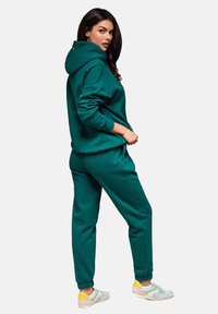 Tearfärgad hoodie och joggers-set, mjukt material, lös passform, ribbade ärmslut och midja, parat med ljusa skor med gula och blå accenter.