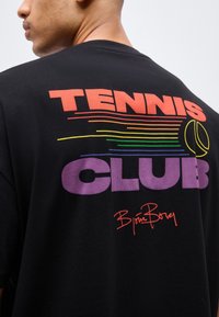 Άνδρας που φοράει μαύρο μπλουζάκι με μεγάλο πολύχρωμο κείμενο «TENNIS CLUB» και ουράνια τόξα μαζί με γραφικό τενιστικής μπάλας στην πλάτη.