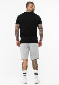 Camiseta negra de cuello redondo, pantalones cortos grises con bolsillo trasero, calcetines blancos con rayas negras y zapatillas blancas. Mostrando brazos y piernas tatuados.