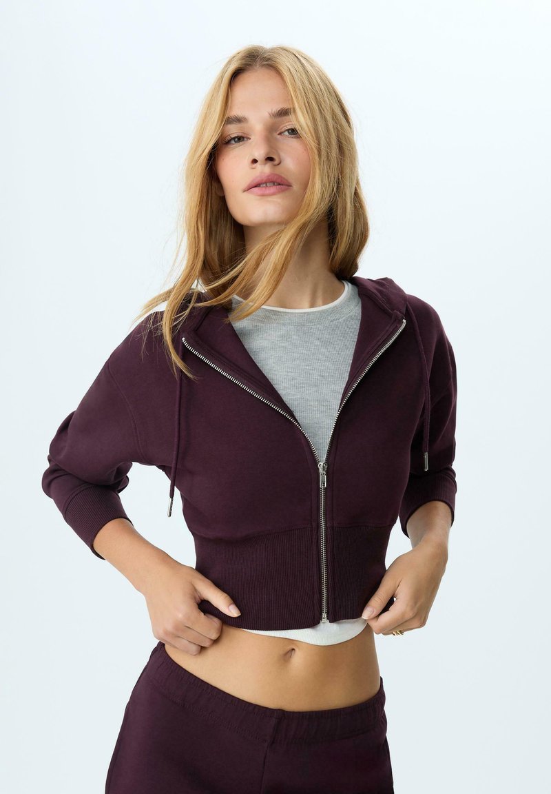 Felpa corta bordeaux con zip, maniche a coste e una maglietta grigia a strati sotto; presenta un cappuccio e una texture liscia.