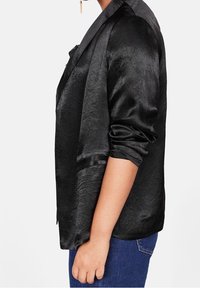 Blazer in satin nero con una texture lucida, dotato di colletto a rever, maniche arrotolate e vestibilità comoda, abbinato a jeans blu scuro.