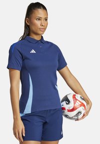 Polo di colore blu navy con accenti azzurri e una chiusura a tre bottoni, abbinato a pantaloni corti blu navy. Tiene in mano un pallone da calcio multicolore.