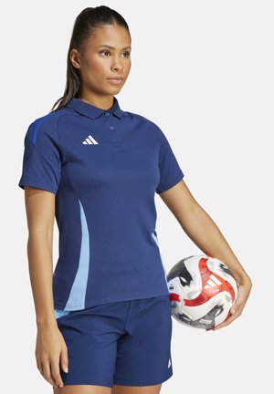 Polo bleu marine avec des accents bleu clair et un col à trois boutons, associé à un short bleu marine. Tient un ballon de football multicolore.