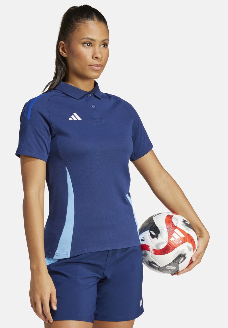 Dunkelblaues Polohemd mit hellblauen Akzenten und einer Dreiknopfleiste, kombiniert mit dunkelblauen Shorts. Hält einen bunten Fußball.