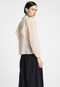 Hellbeige Bluse mit gerafftem Spitzenbesatz, langen Ärmeln und hohem Kragen, kombiniert mit einem schwarzen, fließenden Rock. Glatte, durchsichtige Textur.