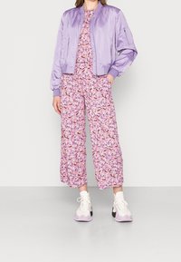Lilac satin bomber jacket with ribbed cuffs over a floral-patterned jumpsuit in shades of pink and purple, paired with white and lilac sneakers.