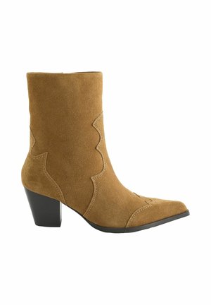 Halve hoge beige suède enkellaars met een spitse neus en decoratieve stiksels, voorzien van een zwarte blokhak en zwarte zool.
