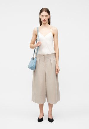 Moteris vilki baltą palaidinę be rankovių, smėlio spalvos plačias kelnes, juodus smailėtus batus ir laiko šviesiai mėlyną peties krepšį priešais paprastą foną.