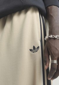 Pantalon beige Adidas avec des bandes noires et un logo, porté par une personne à la peau foncée montrant un bracelet chaîne en argent et une bague à motifs.