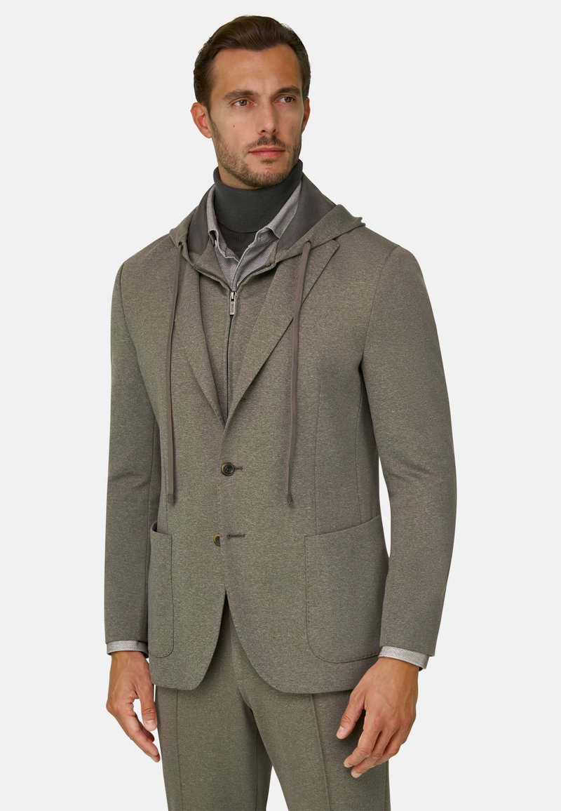 Homme portant une veste de costume taupe avec des poches avant sur un sweat à capuche zippé assorti et un col roulé foncé, debout devant un fond uni.
