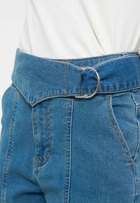 Pantalon en denim bleu taille haute avec une large ceinture maintenue par une boucle double anneau en D argentée, porté avec une chemise blanche rentrée.
