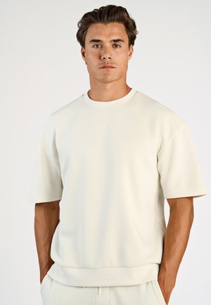 Junger Mann mit mittelbraunen Haaren, der ein schlichtes, übergroßes weißes Kurzarmshirt trägt und die Hände in den Taschen hat, vor weißem Hintergrund.