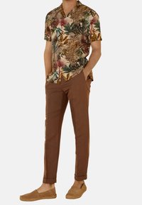 Camicia a maniche corte con colletto e un motivo di foglie colorate; pantaloni slim-fit marroni; espadrillas color nocciola; le trame dei tessuti variano.
