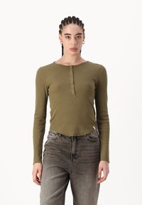 Olivenfarget langermet topp med rund hals og knappelukking. Ribbete tekstur. Kombinert med falmede grå, høyhåndte jeans.