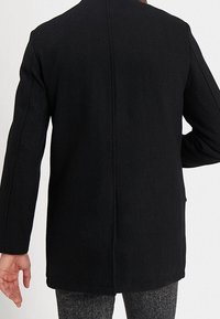 Černý tkaný kabát s rovnou siluetou. Má dlouhé rukávy, vysoký límec a středový šev na zádech. Minimalistický design.