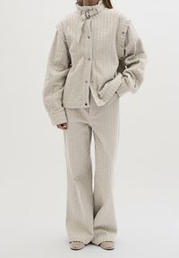 Henkilö, jolla on päällään kermaisen värinen teksturoitu takki ja siihen sopivat leveälahkeiset housut, joiden metalliset varpaat näkyvät yksinkertaista taustaa vasten.