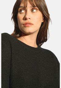 Mustasukkainen neule, jossa on pyöreä kaula-aukko, ominaisuutenaan teksturoitu kangas ja löysä malli. Mallilla on keskipitkä hiustyyli ja neutraali ilme.