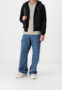 Mężczyzna ubrany w czarną kurtkę na zamek, beżową koszulkę Calvin Klein, niebieskie spodnie cargo, białe trampki, trzymający czarną torebkę na pasku na białym tle.