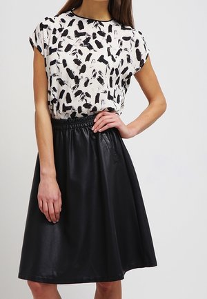 Frau trägt ein weißes kurzärmeliges Oberteil mit schwarzen abstrakten Pinselstrichen und einen schwarzen knieumspielenden Lederminirock mit elastischem Bund.