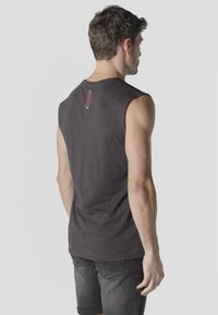 Hombre con camiseta sin mangas gris oscuro y pantalones cortos de mezclilla negros remangados, de pie con la espalda hacia la cámara contra un fondo liso.