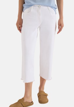 Femme portant un jean blanc raccourci avec des ourlets effilochés, un haut bleu clair et des chaussures marron à enfiler sur fond blanc.