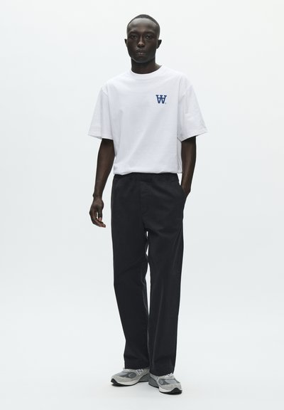 T-shirt blanc à manches courtes avec un logo bleu marine sur la poitrine, associé à un pantalon noir et des baskets grises. Design simple avec une coupe décontractée.