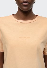 Brzoskwiniowa koszulka z okrągłym dekoltem, krótkimi rękawami i subtelnym nadrukiem logo na piersi. Miękki, lekki materiał o gładkiej fakturze.