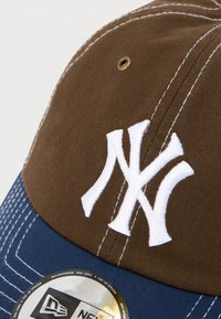 Καπέλο μπέιζμπολ με καφέ κορυφή, μπλε γείσο και λευκό κεντημένο λογότυπο. Διαθέτει λευκή ραφή και κυκλικό ετικέτα μάρκας στο γείσο.