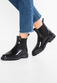 Botas de tobillo negras y brillantes con paneles laterales elásticos, puntera redondeada y suela de goma texturizada, combinadas con jeans de mezclilla azul cortos.