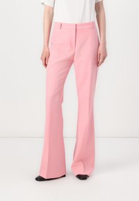 Różowe szyte na miarę spodnie rozwijane, wykonane z gładkiego materiału. Charakteryzują się średnim stanem, bocznymi kieszeniami i schludnym wykończeniem. U dołu widać czarne buty.