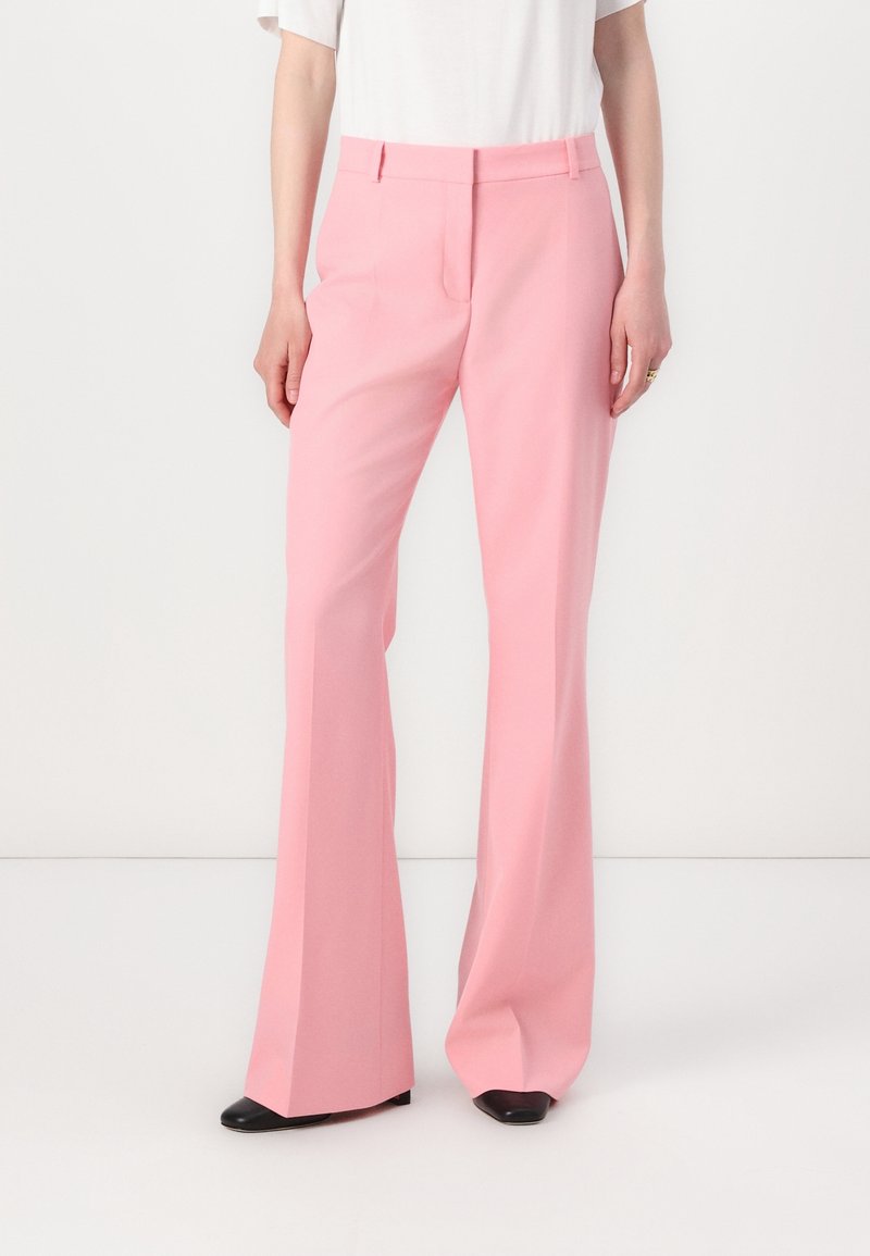 Różowe szyte na miarę spodnie rozwijane, wykonane z gładkiego materiału. Charakteryzują się średnim stanem, bocznymi kieszeniami i schludnym wykończeniem. U dołu widać czarne buty.