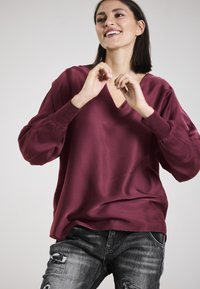 Burgundy, V-neck blouse with balloon sleeves, made of smooth fabric. Paired with distressed gray jeans showing rips and fraying details.