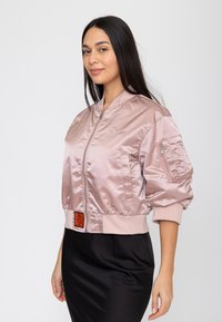 Donna con capelli lunghi e scuri che indossa una giacca bomber lucida rosa chiaro e una gonna nera, in piedi davanti a uno sfondo semplice.