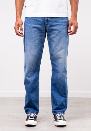 Ein Mann trägt blaue Jeans mit geradem Bein, schwarz-weiße Sneaker und ein weißes T-Shirt und steht auf einem Betonboden vor einer schlichten Wand.