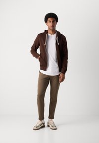 Sudadera marrón con cremallera y cordones blancos, llevada sobre una camiseta blanca y combinada con pantalones ajustados beige y zapatillas beige con detalles verdes.