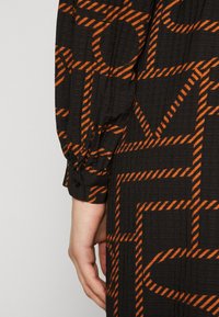 Svart stoffkjole med et geometrisk oransje mønster. Har mansjetter med knapper og teksturert overflate. Detaljert med kraftige linjer og former.