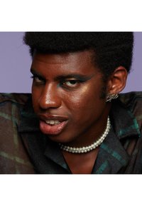 Un portrait rapproché d'une personne à la peau foncée, portant une chemise à carreaux noire transparente, un collier de perles et un maquillage des yeux audacieux avec des accents teal.