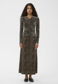 Junge Frau mit lockigem Haar, die ein langes, gerafftes Leopardenmuster-Kleid und schwarze Ballettschuhe trägt, steht vor einem schlichten weißen Hintergrund.