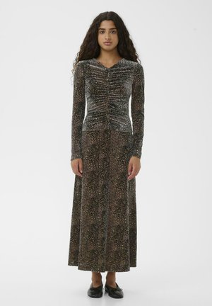 Junge Frau mit lockigem Haar, die ein langes, gerafftes Leopardenmuster-Kleid und schwarze Ballettschuhe trägt, steht vor einem schlichten weißen Hintergrund.