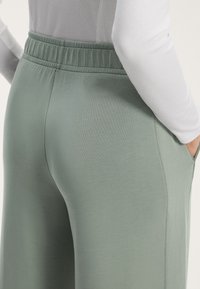 Nära bild av en person som bär ljusgröna byxor med elastisk midja och ribbade sidopaneler, samt en vit långärmad tröja, med handen i fickan.